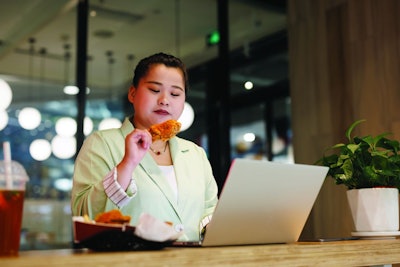 A plump young asian woman, dressed in a green suit, eats hamburgers while working on a laptop in a fast food restaurant.