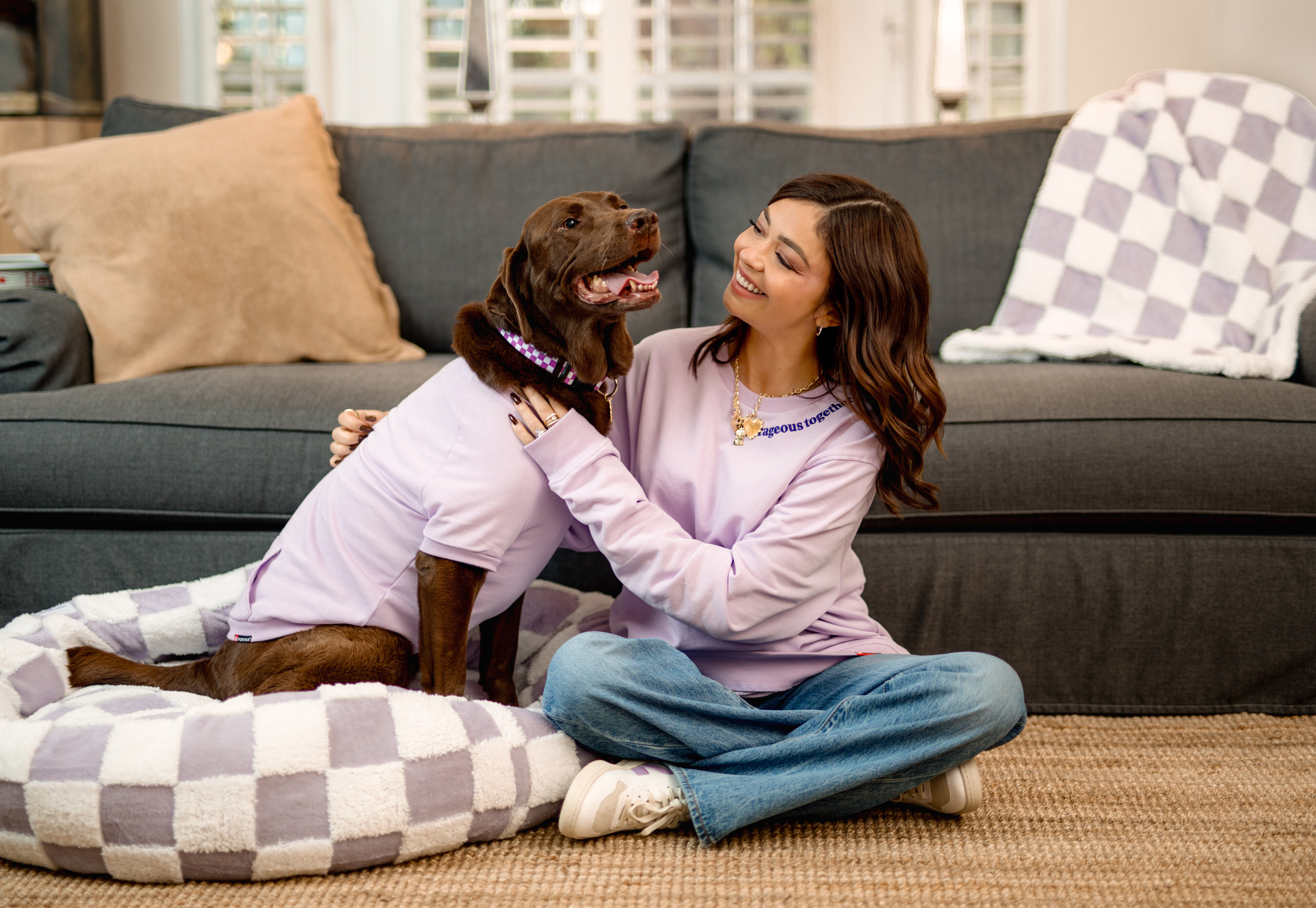 Smiling woman in lavender sweatshirt hugging chocolate Labrador on cozy living room floor