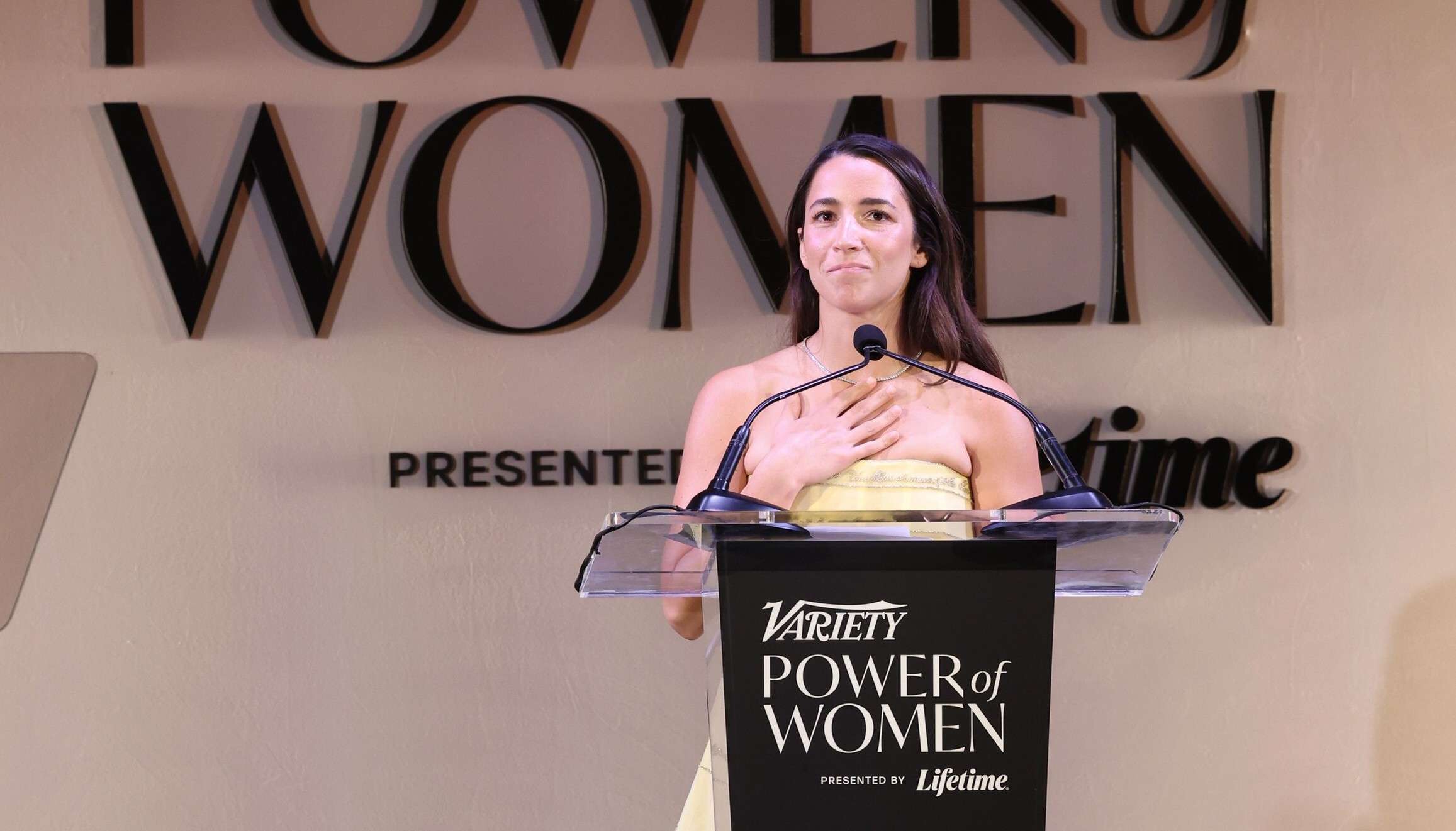 Woman speaking at Variety Power of Women event, standing at podium with large text backdrop