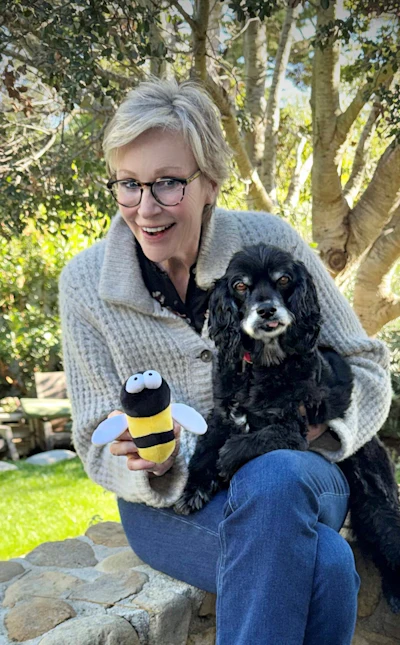 Smiling woman in gray sweater holding black cocker spaniel and yellow bee plush