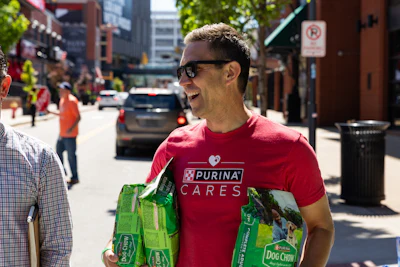 Smiling person in Purina Cares red shirt carrying Dog Chow pet food bags on city street