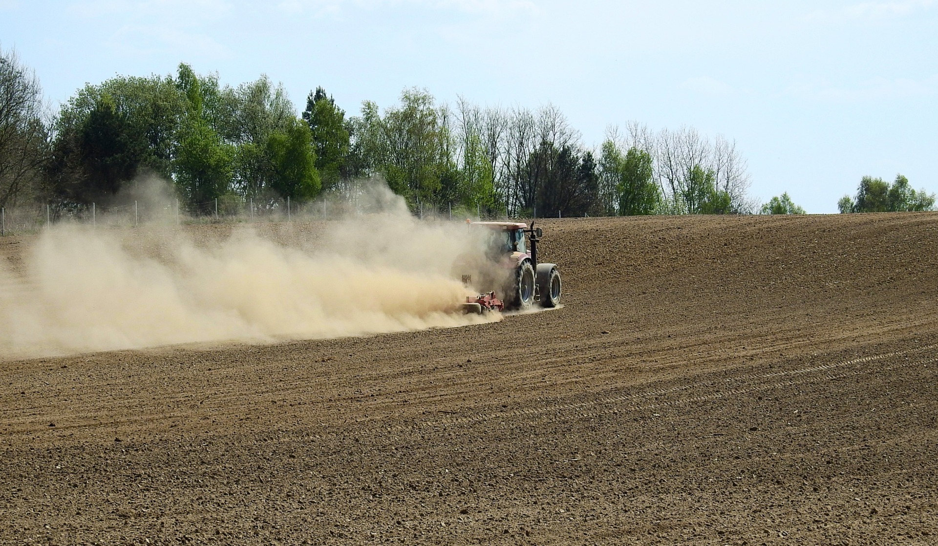 Tractor kicking up dust while plowing brown agricultural field with trees in background