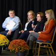Four-person panel discussion on stage with yellow and orange mum flower arrangements