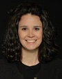 Professional headshot of a smiling woman with dark curly hair against a dark background
