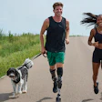 Two runners jogging with dogs on a paved path surrounded by green fields