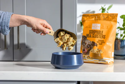 Hand scooping dog food clusters from a measuring cup into a blue pet bowl with packaging visible