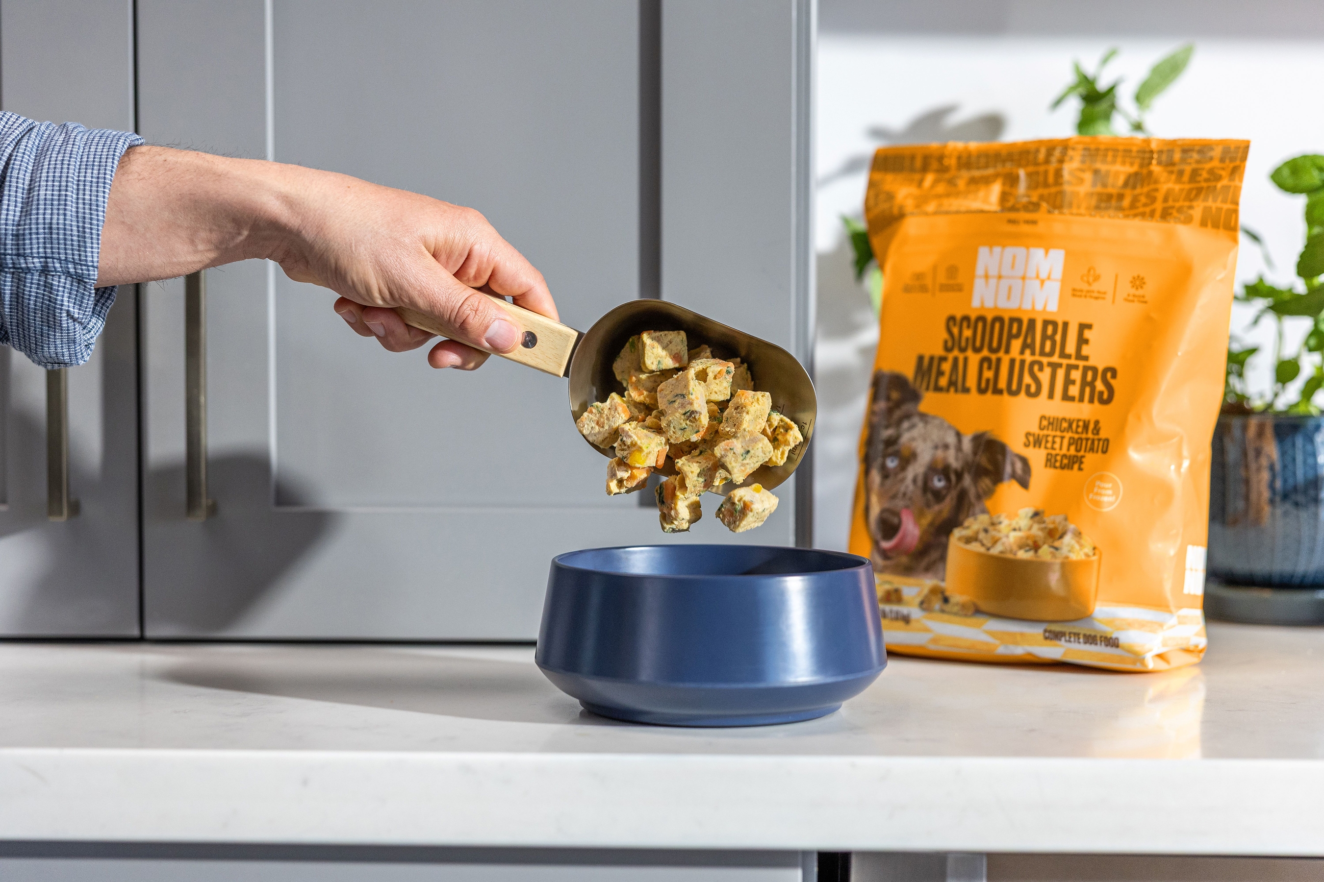 Hand scooping dog food clusters from a blue bowl next to orange pet food packaging