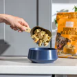 Hand scooping dog food clusters from a blue bowl next to orange pet food packaging