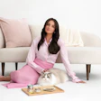 Woman in pink outfit sitting with white cat on cream sofa, pet food products on wooden tray