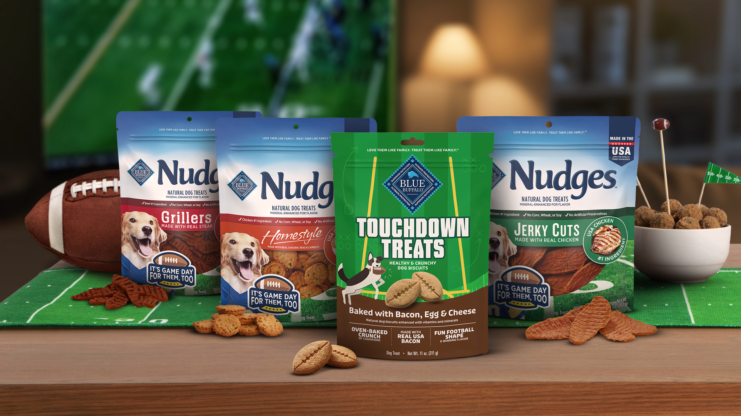 Nudges dog treats product lineup displayed on green football surface with football and accessories