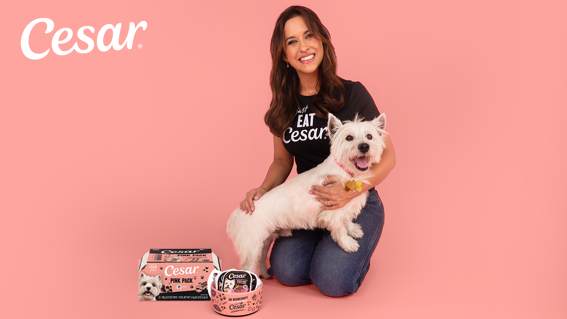 Woman holding white dog wearing Cesar branded shirt with pet food products on pink background