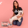 Woman holding white dog wearing Cesar branded shirt with pet food products on pink background