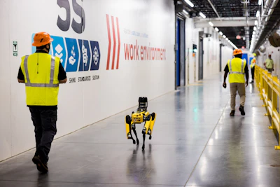 Workers in safety vests and hard hats supervise a yellow robotic device in an industrial warehouse facility with corporate logos on the wall.