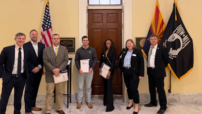 Six professionals stand together in formal attire before flags in an official government building during a recognition event.