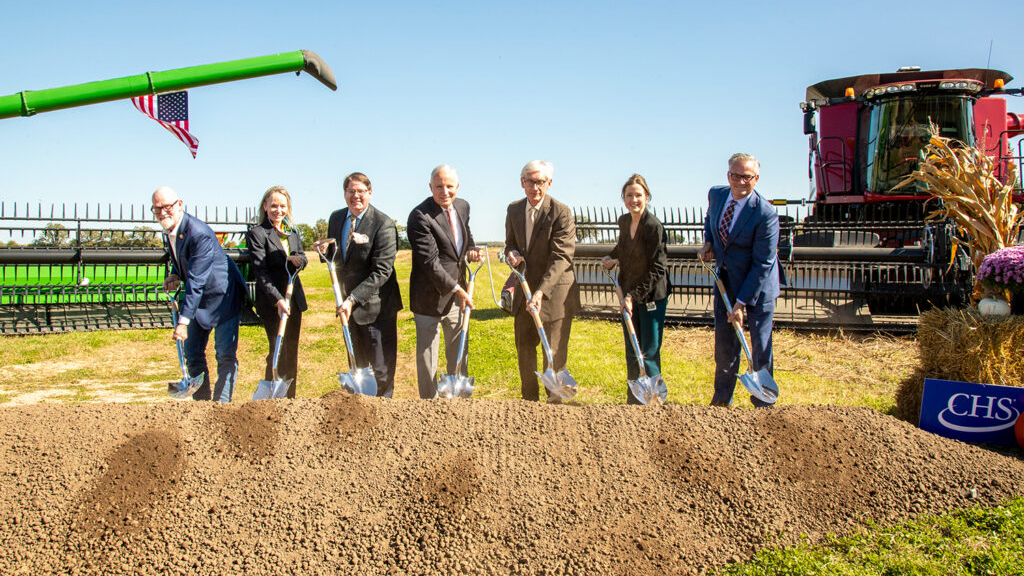 Group of seven business professionals holding shovels at a groundbreaking ceremony with green farm equipment and CHS signage