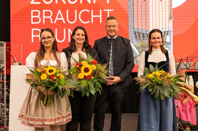 Four people in traditional Bavarian attire holding colorful flower bouquets at a cultural event with orange promotional backdrop