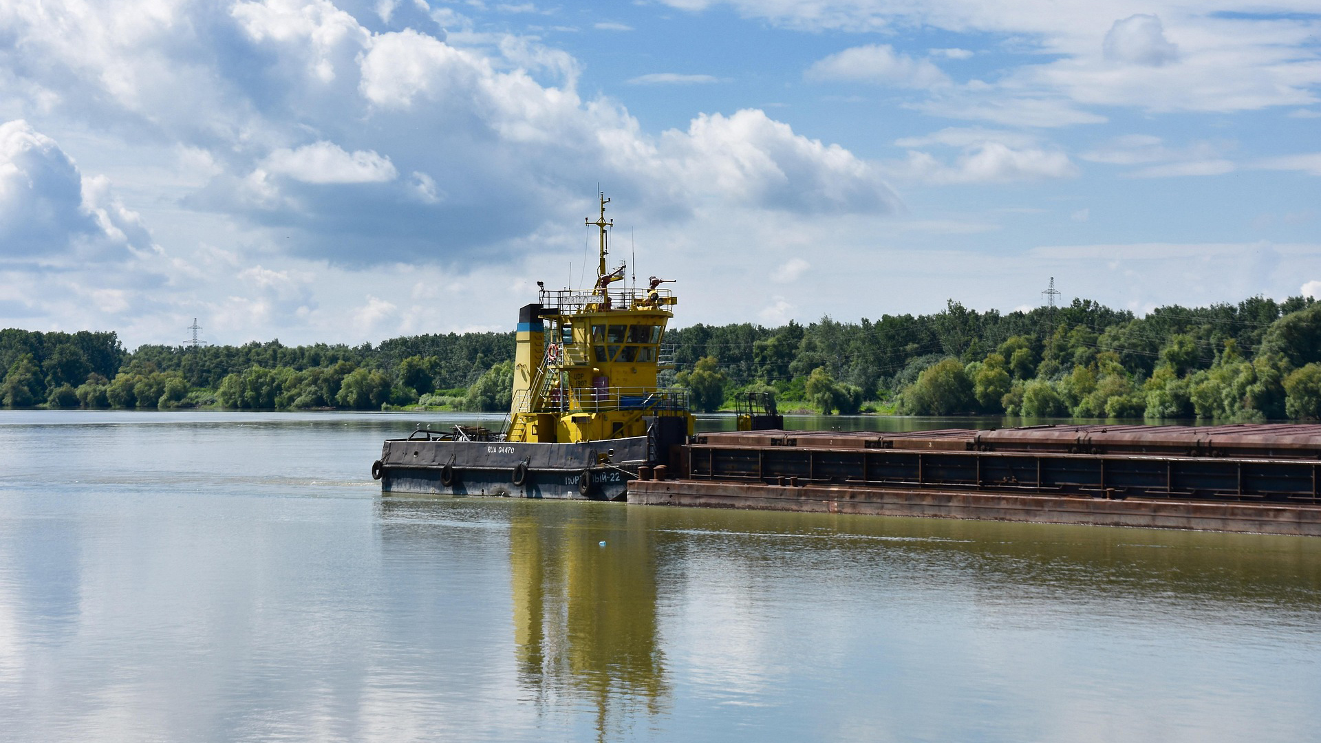 Yellow tugboat pushing a loaded barge on a river with forested banks under blue sky