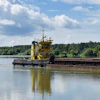 Yellow tugboat pushing a loaded barge on a river with forested banks under blue sky