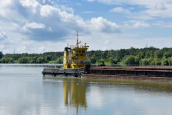 Yellow tugboat pushing a loaded barge on a river with forested banks under blue sky
