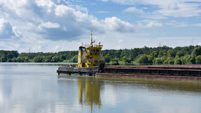 Yellow tugboat pushing a loaded barge on a river with forested banks under blue sky