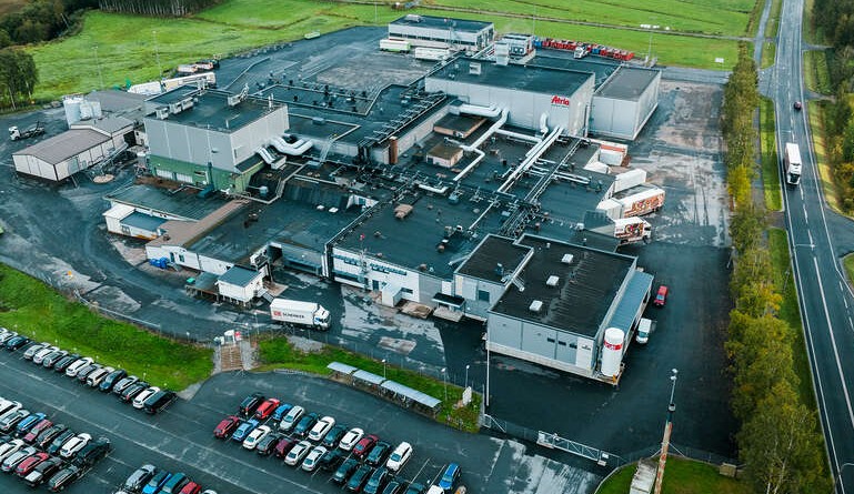 Aerial view of large industrial manufacturing facility with multiple buildings, parking lots, and surrounding green fields