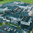 Aerial view of large industrial manufacturing facility with multiple buildings, parking lots, and surrounding green fields