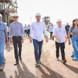 Group of officials in hard hats touring a waterfront industrial construction site with equipment and workers visible