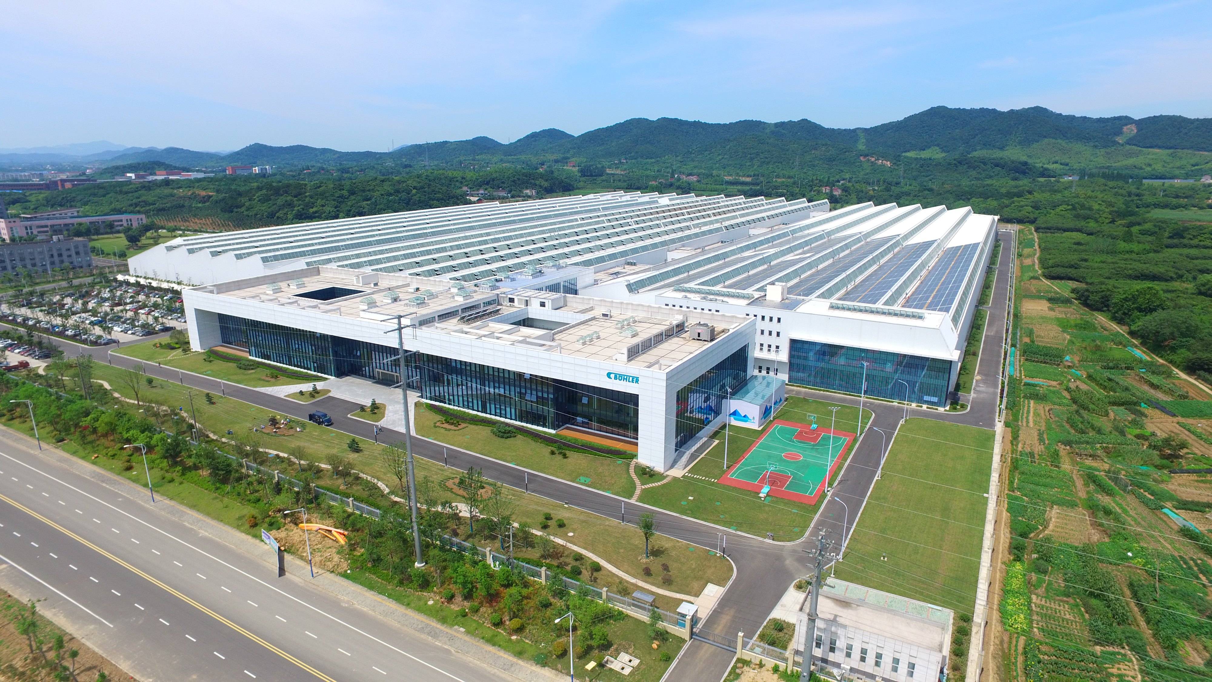 Aerial view of modern industrial facility with solar panel roof, sports courts, and surrounding green fields