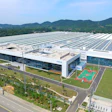 Aerial view of modern industrial facility with solar panel roof, sports courts, and surrounding green fields