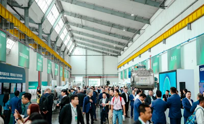 Large industrial facility with yellow overhead cranes, crowds of attendees in business attire, and green branded signage panels on walls.