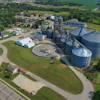 Aerial view of grain elevator facility with blue cylindrical silos, processing equipment, and surrounding agricultural fields