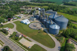 Aerial view of grain elevator facility with blue cylindrical silos, processing equipment, and surrounding agricultural fields