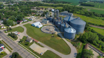 Aerial view of grain elevator facility with blue cylindrical silos, processing equipment, and surrounding agricultural fields