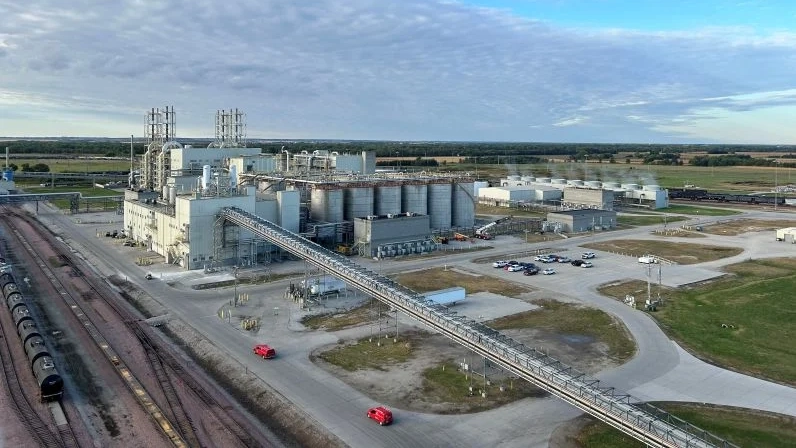 Large grain elevator facility with cylindrical storage silos, industrial buildings, and railroad tracks in rural landscape