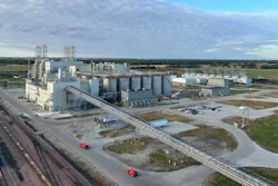Large grain elevator facility with cylindrical storage silos, industrial buildings, and railroad tracks in rural landscape