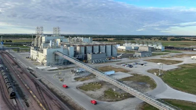 Large grain elevator facility with cylindrical storage silos, industrial buildings, and railroad tracks in rural landscape