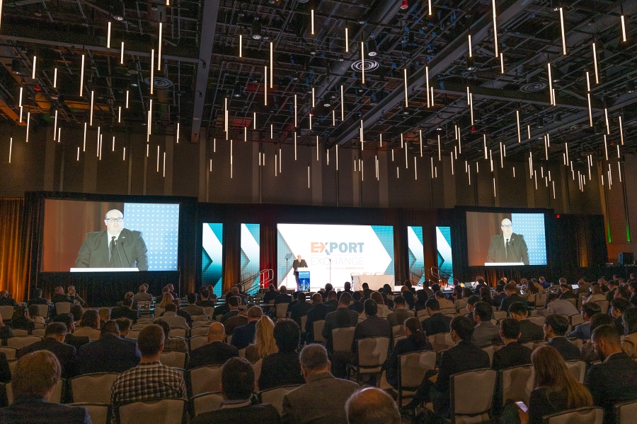Large conference hall with hundreds of attendees seated facing multiple projection screens displaying an export-themed business presentation with pendant lighting overhead.
