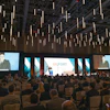Large conference hall with hundreds of attendees seated facing multiple projection screens displaying an export-themed business presentation with pendant lighting overhead.