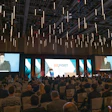 Large conference hall with hundreds of attendees seated facing multiple projection screens displaying an export-themed business presentation with pendant lighting overhead.