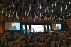 Large conference hall with hundreds of attendees seated facing multiple projection screens displaying an export-themed business presentation with pendant lighting overhead.