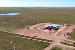Aerial view of rural construction site with red barn and blue roof surrounded by green fields and dirt roads