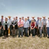 Group of professionals wearing hard hats and business attire posing together in front of yellow construction excavators at an outdoor event