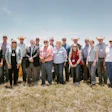 Group of professionals wearing hard hats and business attire posing together in front of yellow construction excavators at an outdoor event