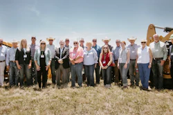 Group of professionals wearing hard hats and business attire posing together in front of yellow construction excavators at an outdoor event