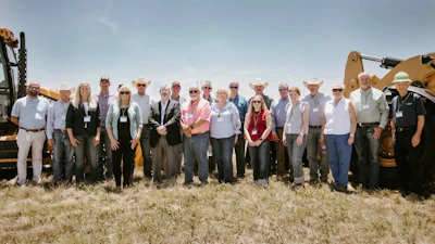 Group of professionals wearing hard hats and business attire posing together in front of yellow construction excavators at an outdoor event
