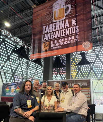 Group of six people posing at a trade show booth beneath a large 'Taberna de Lanzamientos' beer sign sponsored by U.S. Grains & Bioproducts Council