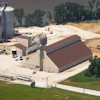 Aerial view of grain processing facility with metal silos, storage buildings, and railroad tracks surrounded by fields