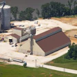 Aerial view of grain processing facility with metal silos, storage buildings, and railroad tracks surrounded by fields