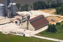 Aerial view of grain processing facility with metal silos, storage buildings, and railroad tracks surrounded by fields