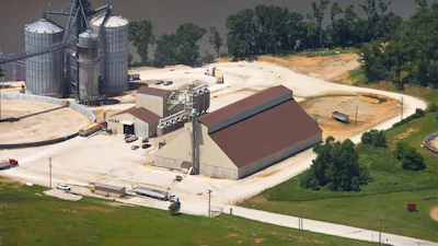 Aerial view of grain processing facility with metal silos, storage buildings, and railroad tracks surrounded by fields
